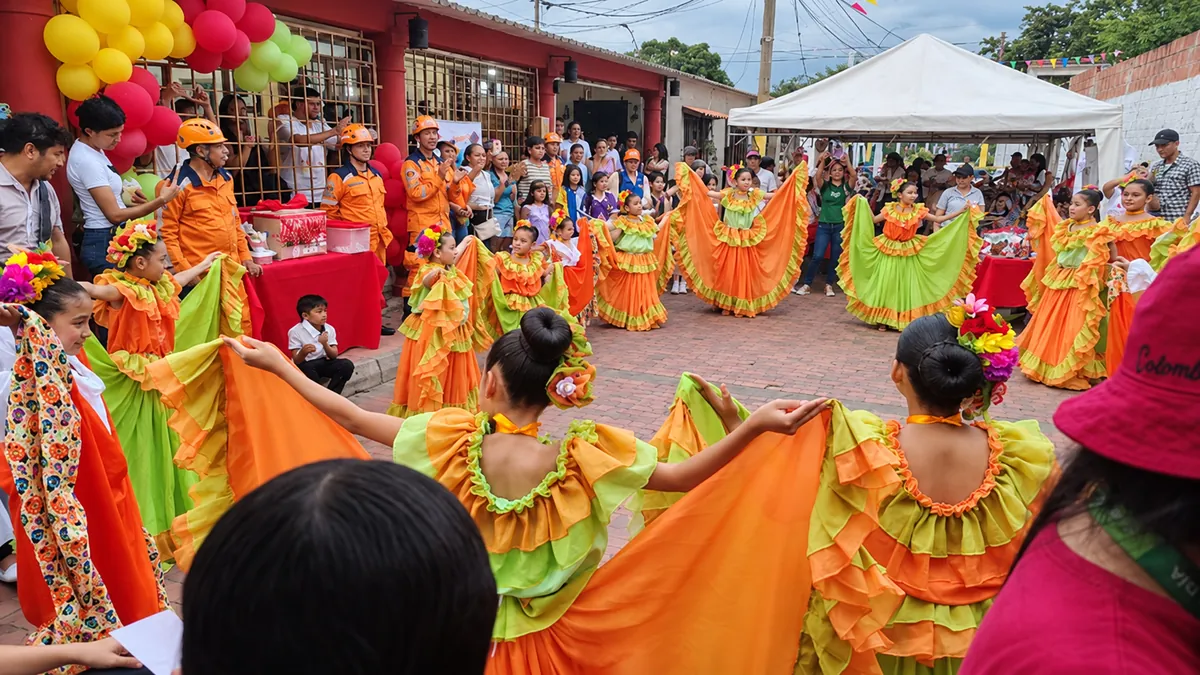 Niñas con trajes típicos naranja y verde bailan en ronda en una calle adoquinada rodeadas de público